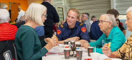 Foto van een brandweerman aan tafel met meerdere ouderen tijdens een lunch